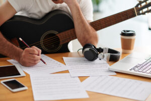 Homem com violão no colo apoiado em uma mesa enquanto escreve.